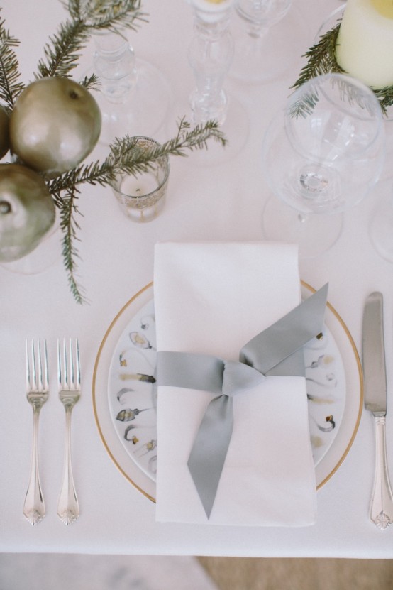 a beautiful neutral NYE table setting with a white charger and a printed plate, evergreens and pillar candles, silver cutlery