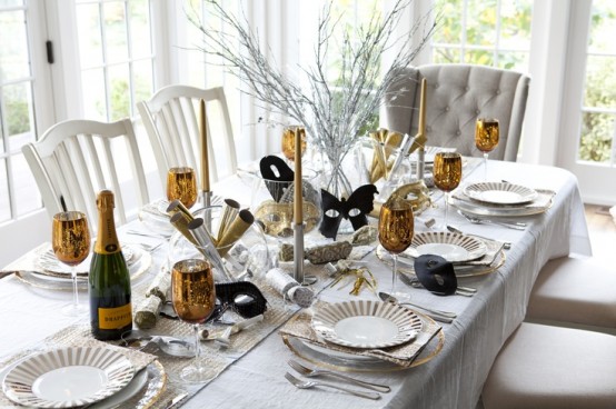 a white NYE tablescape with a silver table runner, white plates, gold glasses, gold candles and masks is an elegant idea