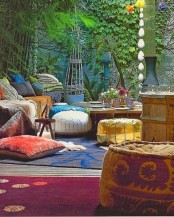 a colorful boho patio with colorful textiles, pillows, rugs, a gold and a printed ottoman plus greenery
