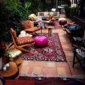 a colorful boho patio done with rugs, printed furniture, pillows, ottomans, a gold ottoman and potted plants