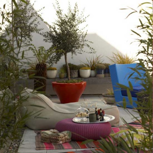 a bright patio with colorful furniture, rugs and ottomans plus lots of potted greenery and blooms