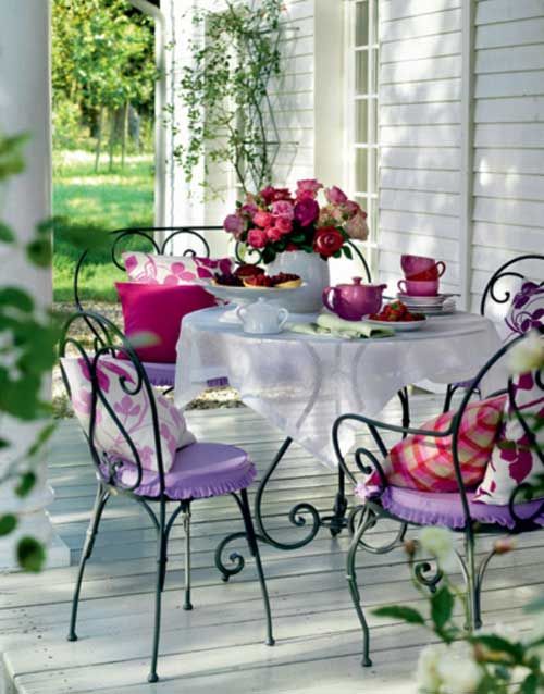 a refined vintage terrace with black forged furniture, lilac and pink pillows, a lace tablecloth and hot pink blooms on the table
