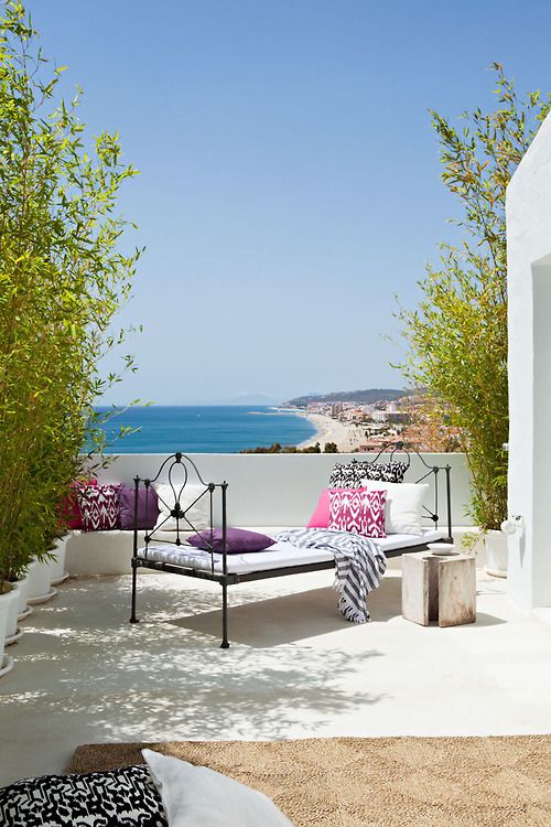 a lovely Mediterranean terrace with a black forged daybed with pink and purple pillows, greenery around and a cool sea view is amazing