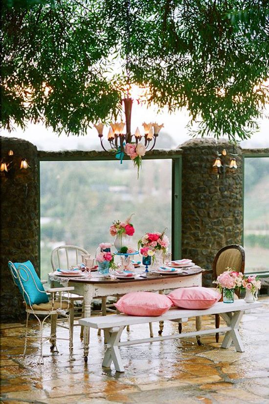 an eclectic shabby chic terrace with a shabby dining table, chairs and benches, pillows, cushions and bold blooms plus a chic chandelier over the zone