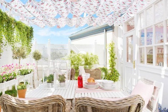 a bright feminine terrace with white planked tables, woven chairs, floral upholstery and a roof and potted plants and blooms around