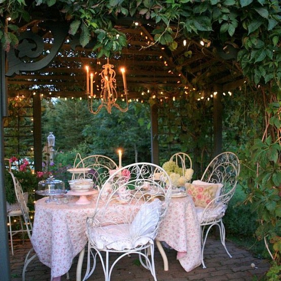 a pastel vintage terrace surrounded with greenery, with a chandelier and lights over the space, a white forged table and chairs, a pink tablecloth and pink pillows is amazing