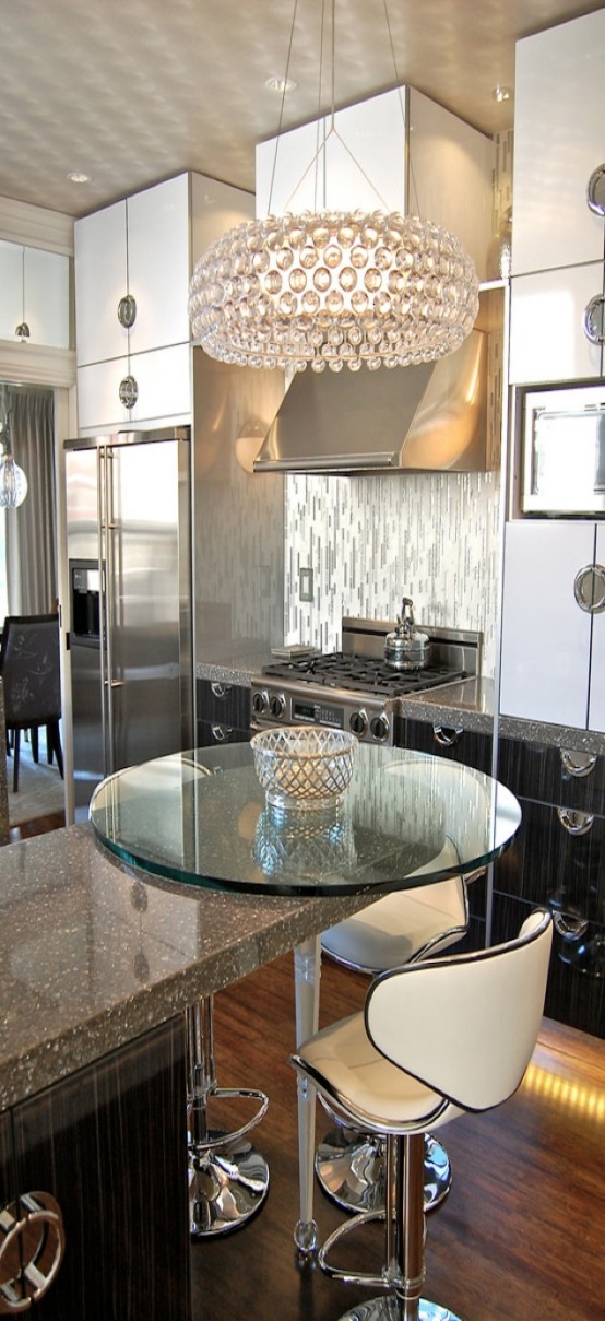 a glam kitchen with dark cabinetry, shiny silver handles, a large crystal chandelier and a glass countertop