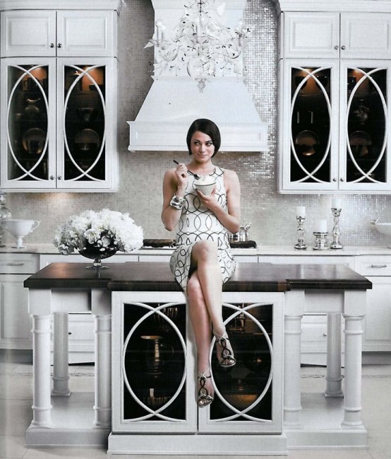 a chic glam ktichen with white furniture, a shiny white tile backsplash, a stylish kitchen island with a black countertop and a crystal chandelier