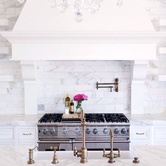 a vintage glam kitchen with white furniture, white marble tiles, vintage brass faucets and a large hood