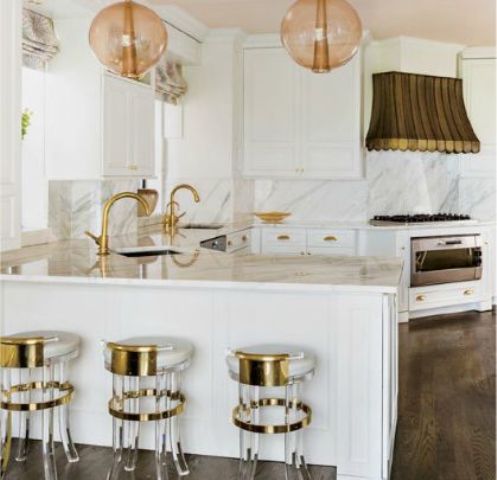 a modern glam kitchen with white furniture, a white marble backsplash and coutnertops, pendant lamps and a hood over the cooker