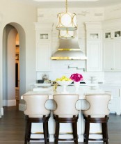a small vintage glam kitchen in white, with elegant cabinetry, white stone countertops, vintage chairs and a shiny metal hood