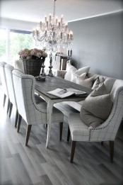 a grey dining room with a vintage whitewashed table, upholstered chairs, a crystal chandelier, a large basket with blooms and cool pillows