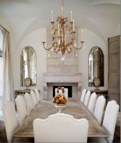 a neutral dining room with a long whitewashed table and neutral chairs, a fireplace, a crystal chandelier and vintage mirrors