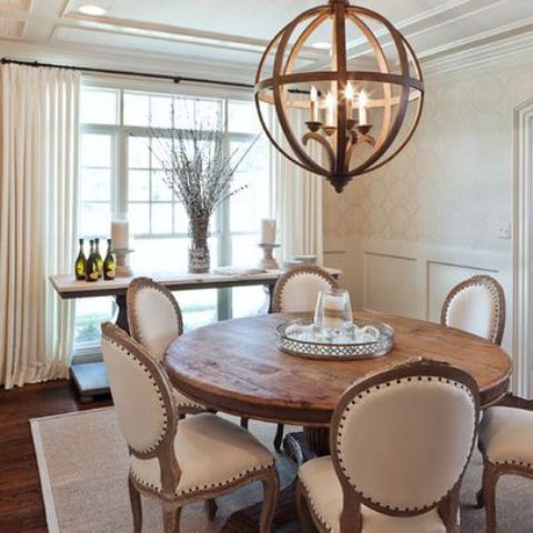 a refined vintage neutral dining room with paneled walls, a round table and neutral chairs, a sphere chandelier and a console with some decor