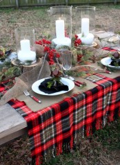 a rustic Christmas tablescape with a plaid runner and burlap placemats, tree stumps with large candles, evergreens and berries and large bells plus black napkins
