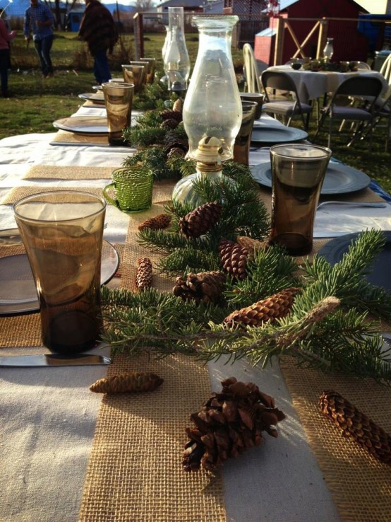 a rustic Christmas tablescape with a burlap table runner and placemats, clear glass chargers and evergreens, pinecones and table lamps
