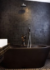 a modern black bathroom clad with small scale tiles, a black oval tub and a black vanity clad with tiles plus a white sink