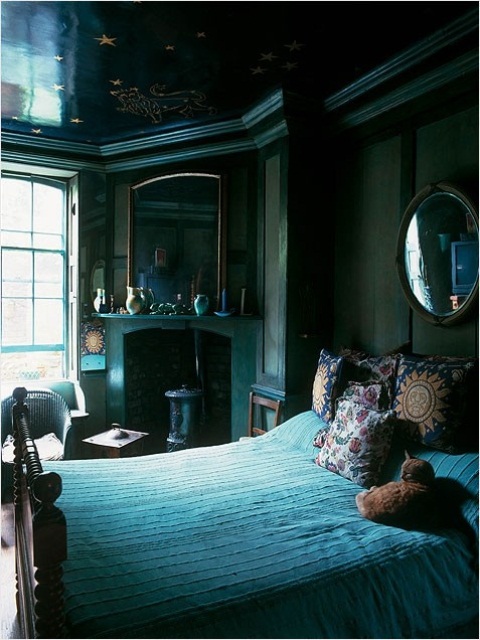 a moody bedroom with dark green walls, a fireplace, a mirror over the mantel, a round mirror over the bed and a couple of chairs