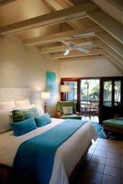 an attic bedroom done in neutrals, with wooden beams on the ceiling, a tiled floor, a bed with turquoise accents and green chairs