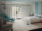 a peaceful neutral bedroom with light blue and turquoise accents, blue stripes on the wall and ceiling, a white bed and bedding, a turquoise lounger and a chic chandelier