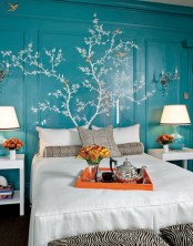 a bold turquoise bedroom with paneling on the walls, with a painted tree with leaves, a bed with neutral and grey bedding, white nightstands and turquoise and white table lamps