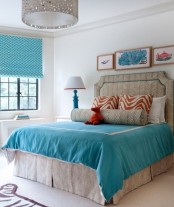 a lively bedroom in neutrals, with an upholstered bed and printed and turquoise bedding, a mini gallery wall, turquoise printed shades and table lamps with turquoise bases