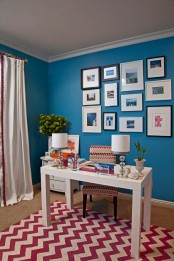 a bold blue home office with a lovely grid gallery wall, a white desk and a printed chair, a red and white chevron rug that gives much color to the space