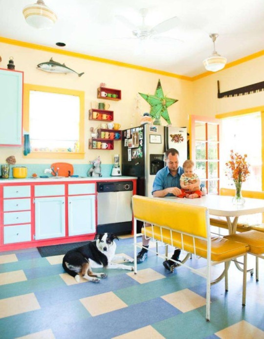 a bright kitchen with yellow walls, powder blue cabinets with pink frames, blue and green tiles, bright yellow furniture