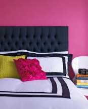 a hot pink bedroom with a black upholstered bed, printed bedding, pink and mustard pillow and a stack of books on the chair is a vivacious space that feels and looks awesome