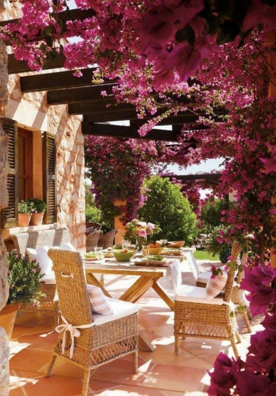 a French chic terrace wiht bougainvillea covering it completely, a wooden table, wicker chairs and neutral textiles