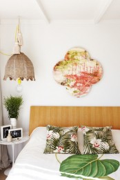 a contemporary tropical bedroom with bright printed pillows, a bold artwork and a wicker lampshade over the bed