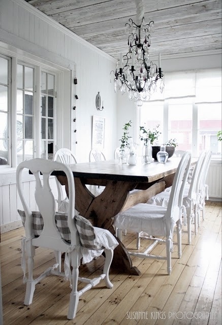 a white dining room in vintage meets rustic style, a large stained table and white chairs, a lovely crystal chandelier and potted greenery