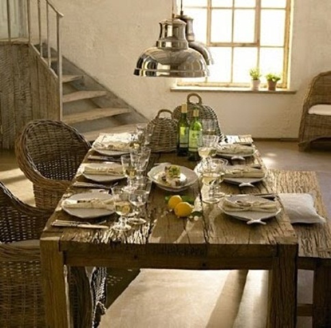 a rustic dining room with an uncovered wooden table, a wooden bench and woven chairs, metal pendant lamps over the space
