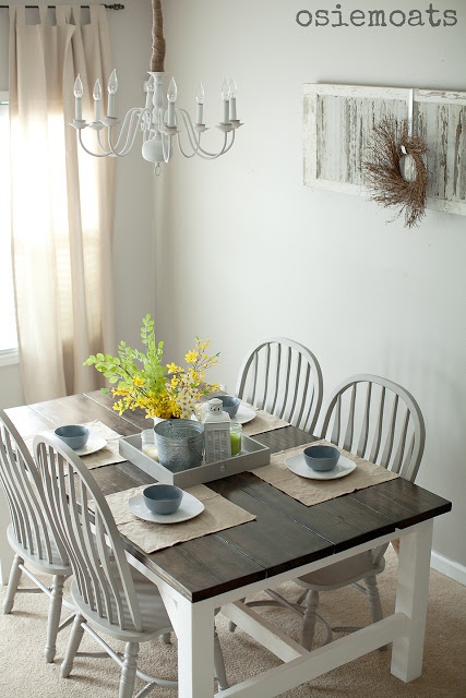 a vintage farmhouse dining space with a black and white dining table, whitewashed chairs, a shabby chic decoration on the wall and a white chandelier