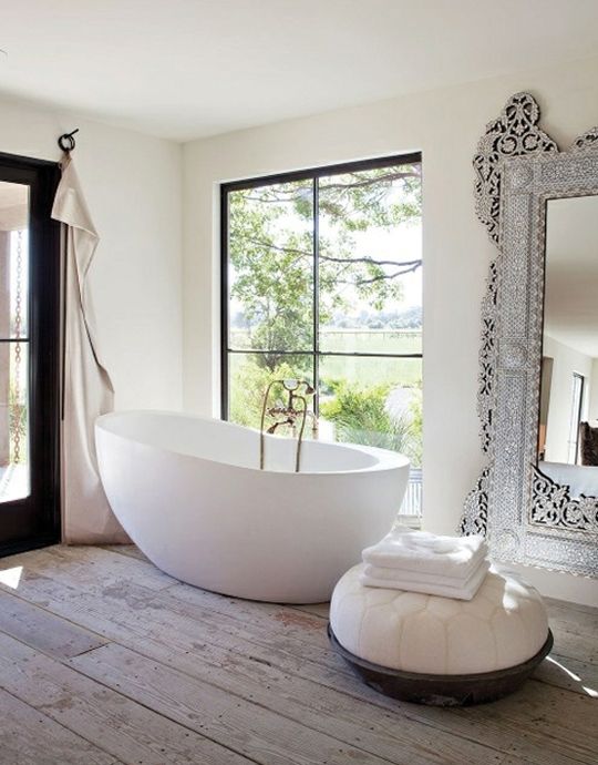 a refined eclectic bathroom with an oval bathtub, an oversized mirror in an ornated frame, a Moroccan ottoman and neutral curtains
