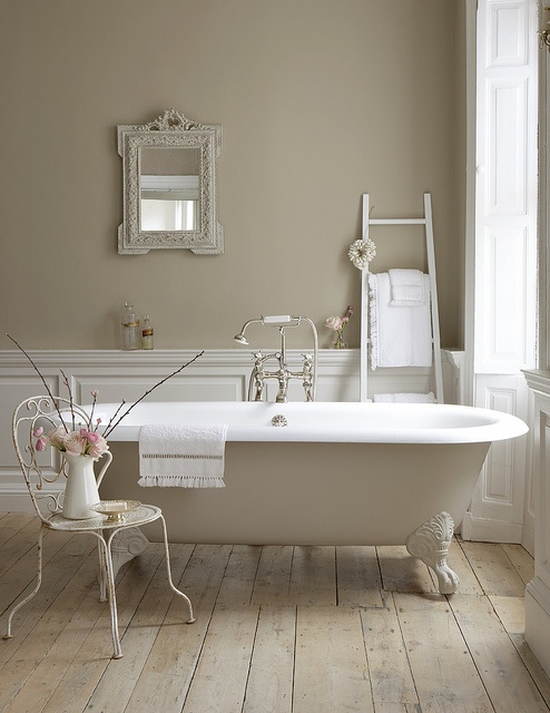 a neutral vintage bathroom with grey walls and white panels, a creamy bathtub, a ladder, a mirror in an ornated frame and a vintage chair