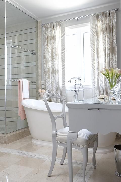a refined neutral bathroom with tan tiles, stripes in the shower space, white furniture and a vanity plus printed floral curtains
