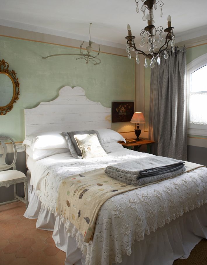 a vintage bedroom with light green walls, a bed with a whitewashed headboard, a nightstand and a chair, a crystal chandelier
