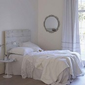 a white shabby chic bedroom with a whitewashed bed with white bedding, a white nightstand and table lamps, a mirror on the wall