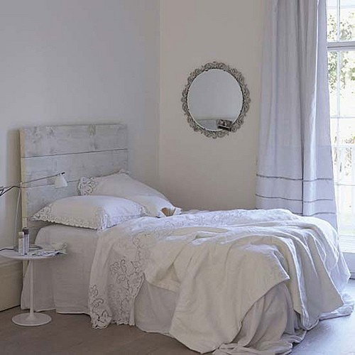 a white shabby chic bedroom with a whitewashed bed with white bedding, a white nightstand and table lamps, a mirror on the wall
