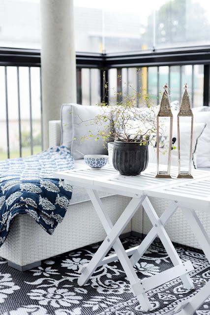a Nordic terrace with a white wicker bench and printed blankets and pillows, white folding tables, potted greenery and candle lanterns