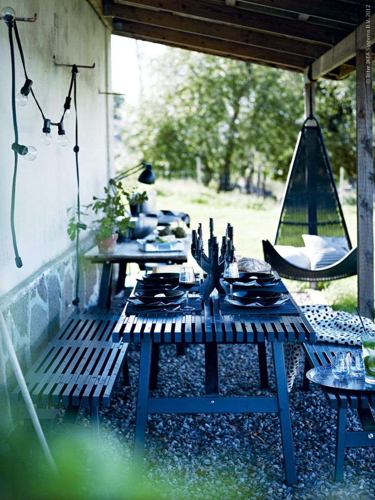 a Scandinavian terrace under a roof, with black garden furniture, a blakc pendant chair, greenery and table lamps and string lights