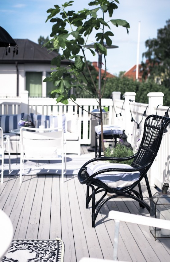 a Scandinavian terrace with a white sofa with striped pillows, white chairs, a black rattan chair with a neutral cushion, potted greenery and lights