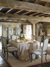 a rustic Provence dining room with stone walls,a  wooden ceiling with beams, a dining table and vintage chairs, pendant lamps and some blooms