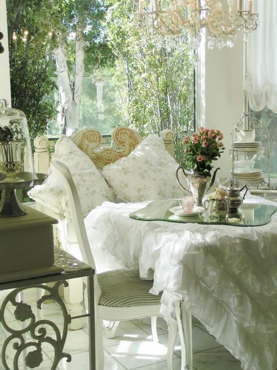 a refined white vintage sunroom with shabby chic and vintage furniture, a crystal chandelier, floral pillows and blooms in vases