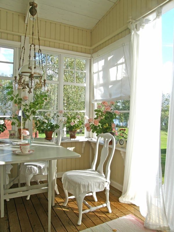 a buttermilk colored vintage sunroom with elegant white furniture, a vintage chandelier and lots of potted blooms on the windowsills