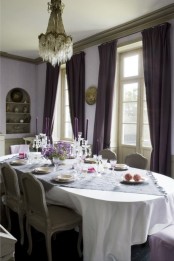 an exquisite French dining room with lilac walls, a curved niche, an oval table and vintage chairs, deep purple curtains and a crystal chandelier