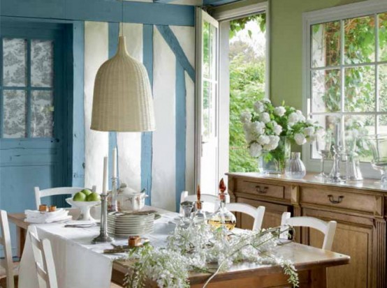 a dreamy French cottage dining room with blue and white walls, a stained sideboard, a table and white vintage chairs, a woven pendant lamp