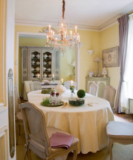 a French chic dining room with pastel yellow walls, a corner cabinet, an oval table and vintage chairs, a crystal chandelier and greenery topiaries