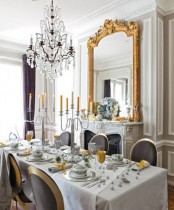 a neutral sophisticated French dining room with a fireplace, a large mirror in a gilded frame, a table and chic grey and gold chairs, a crystal chandelier